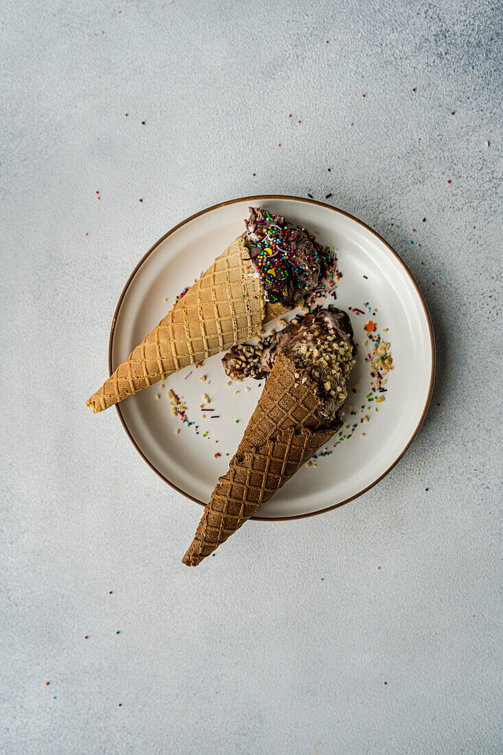 Top view of waffle cones with coffee and chocolate ice cream with multicolored sugar and nuts on a ceramic plate on a concrete background