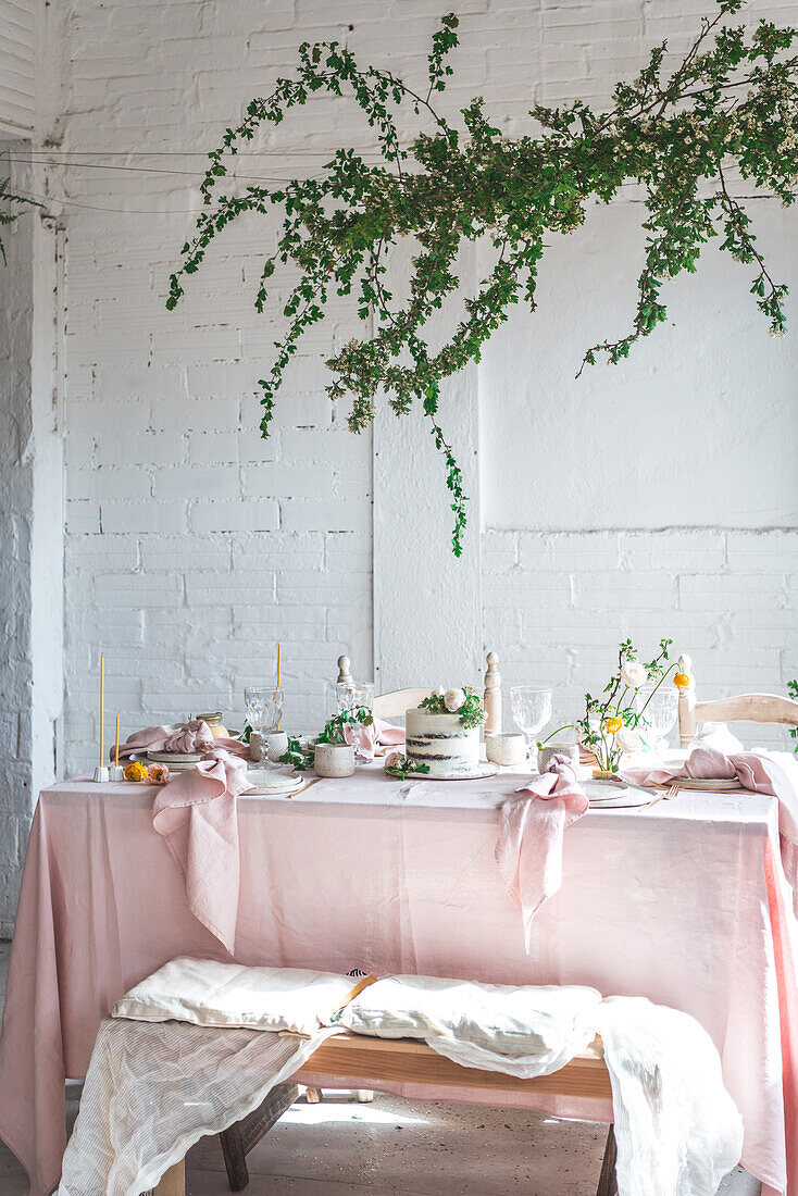 Elegant table served with plates and flowers near yummy cake on pink tablecloth placed near bench covered with cloth against potted plant and white brick wall