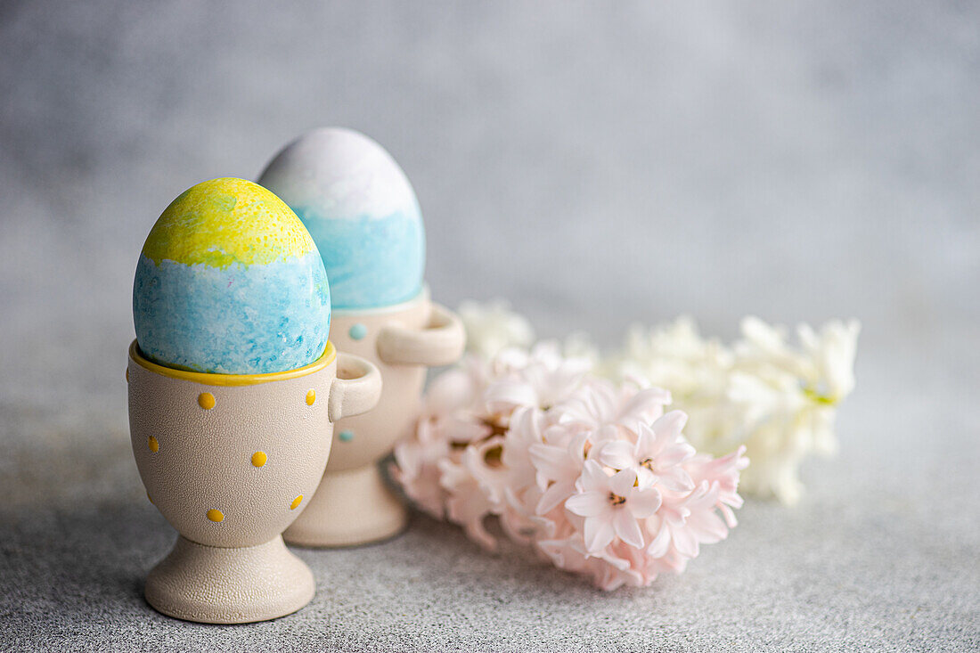 Easter card concept with colorful eggs on eggcups near seasonal flowers on concrete background