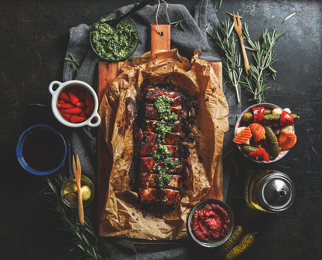 Gebratene BBQ-Rippchen mit hausgemachtem Pesto, eingelegtem Gemüse, Soßen und Rosmarin auf dunklem Beton Küchentisch. Würzig gewürztes Fleisch zum Abendessen. Ansicht von oben