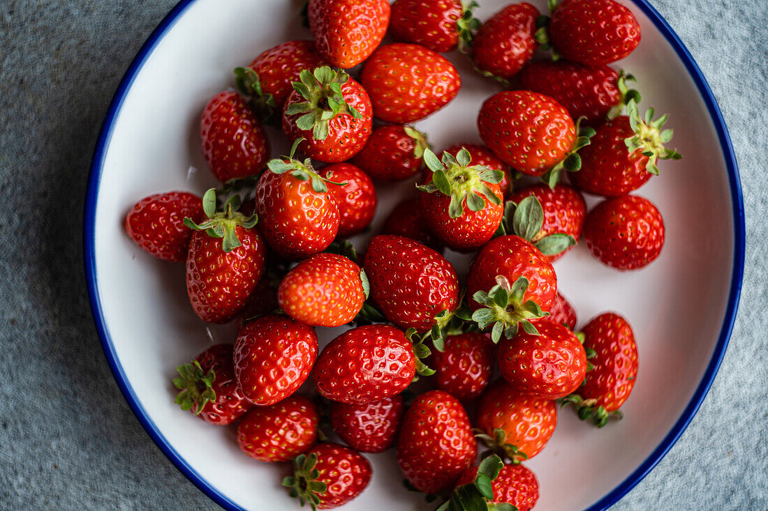 Top view of bright red organic strawberries with green leaves, displayed in a white plate with a blue rim, against a textured gray backdrop.
