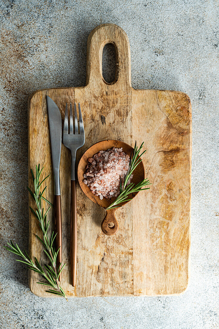 From above rustic kitchen setup on wooden cutting board with a fork, knife, a sprinkle of pink Himalayan salt, fresh rosemary sprigs, laid out on a textured concrete background