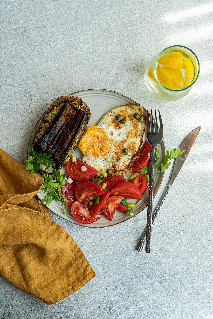 From above of plate with delicious healthy lunch including fried eggs with tomatoes slices, parsley, spring onion, capers served with glass of cold pure water and lemon slices