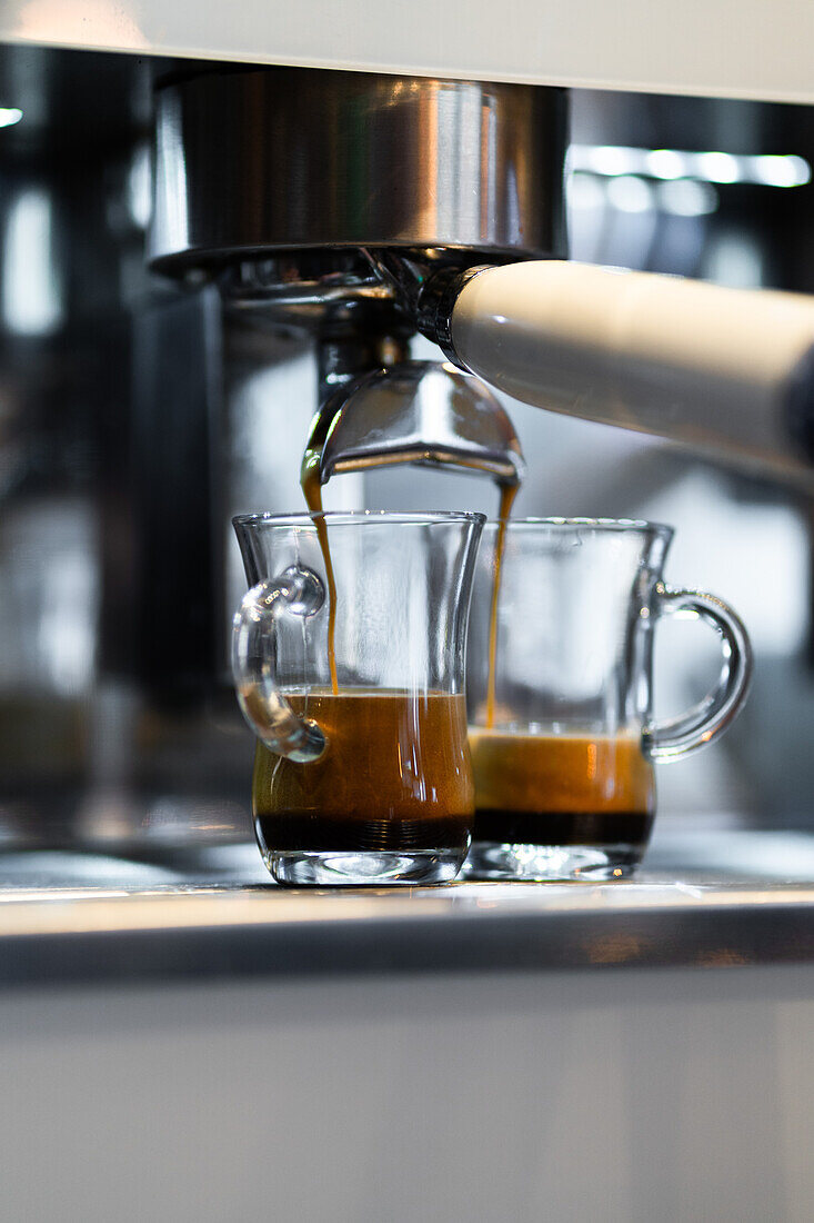 Hot fresh espresso flowing into pair of glass cups from coffeemaker placed on counter