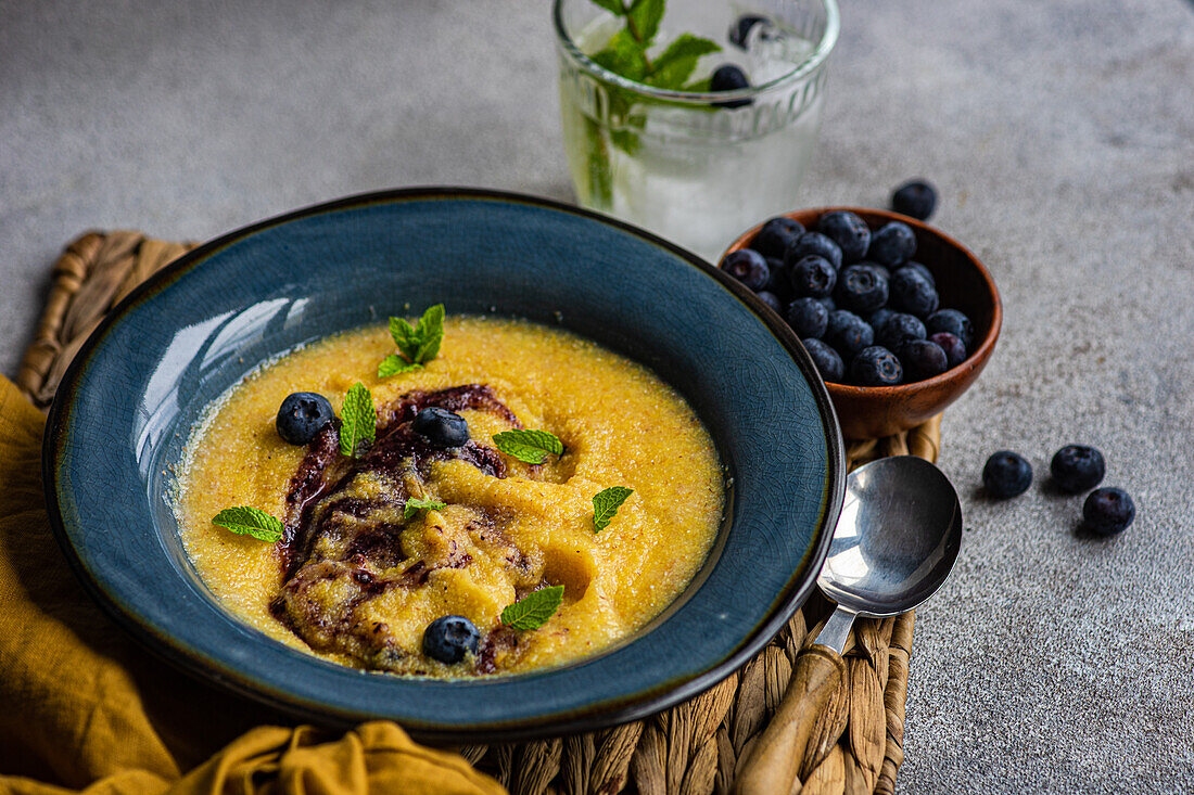 Top view of delicious traditional Italian polenta porridge topped with jam and mint leaves and served on table with bowl of fresh blueberries and glass of cold lemonade during breakfast