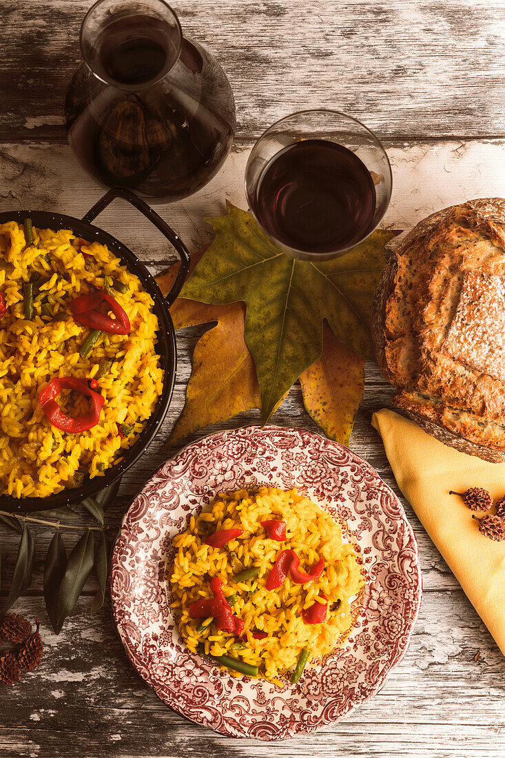 A rustic autumnal table setting featuring paella, artisan bread, and a glass of red wine, showcasing warm, comforting food perfect for the season.