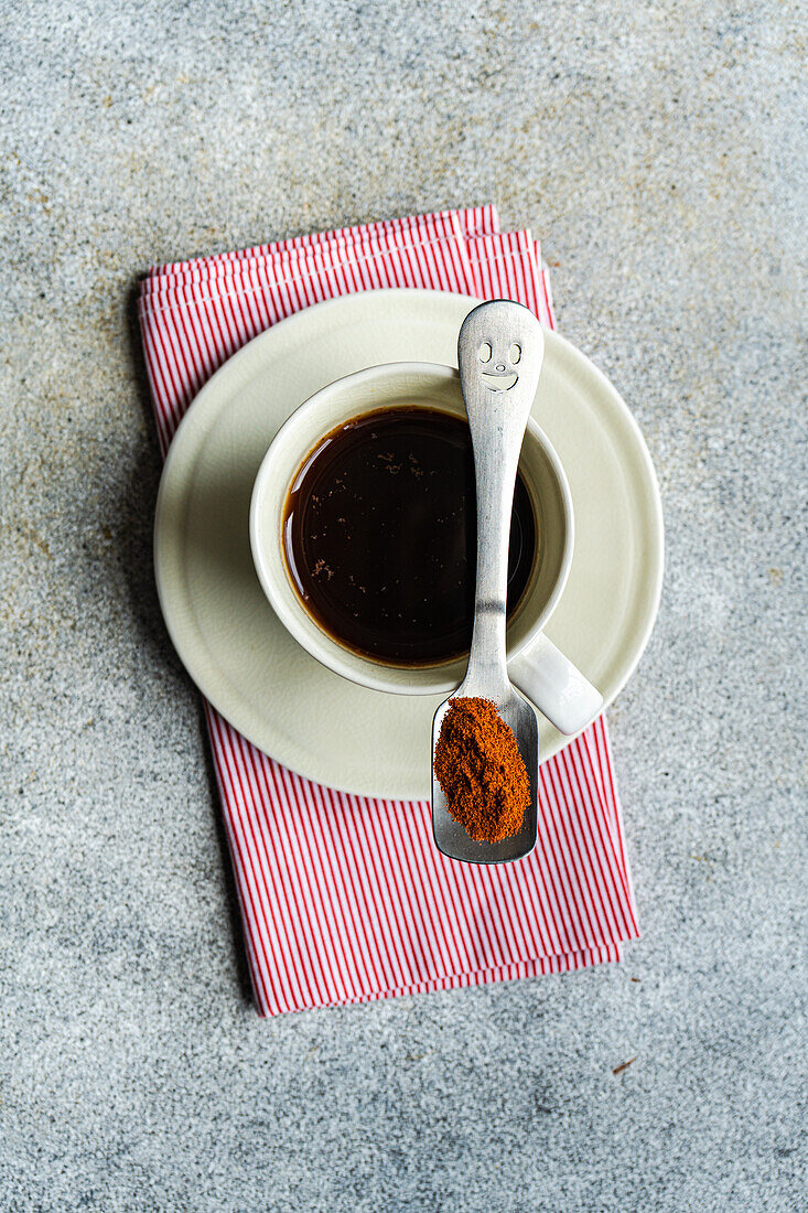 Top view of cup of spicy espresso with spoon with red paprika placed on napkin against gray table