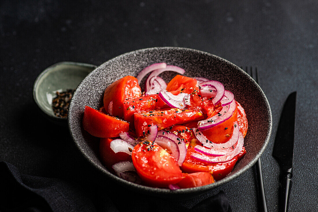 Schüssel mit Salat mit Bio-Tomaten vom Bauernhof und roten Zwiebeln mit Leinsamen und Meersalz auf schwarzem Hintergrund