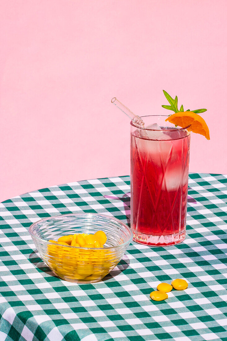 Glass of cold refreshing red cocktail with ice and straw served with orange near transparent bowl with lupin bean on table with checkered tablecloth