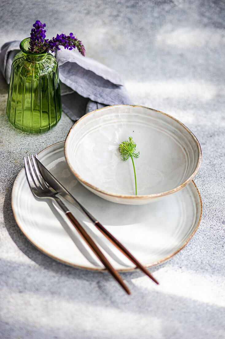 High angle of autumnal table setting with ceramic bowl and plate with flower near fork, knife, napkin and vase with lavender flowers against gray surface