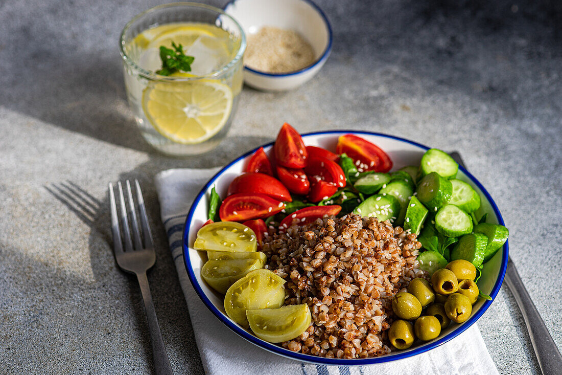 High angle of healthy lunch bowl with boiled organic buckwheat, fresh cucumber and tomato, and fermented tomato and olives served with glass of pure water with lemon, ice and mint against blurred gray surface
