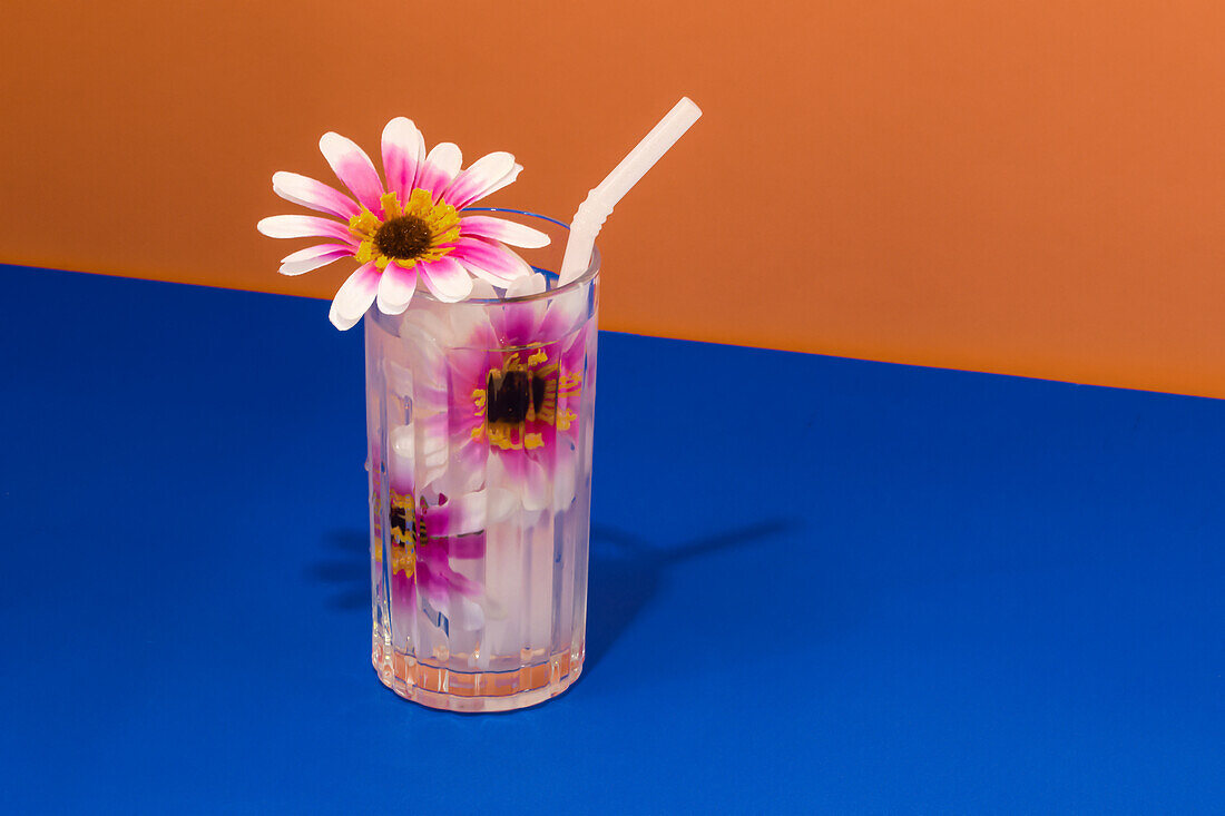 Transparentes Glas mit erfrischendem Kaltgetränk, verziert mit rosa Blumen und Strohhalm auf blauer Fläche vor leuchtend oranger Wand