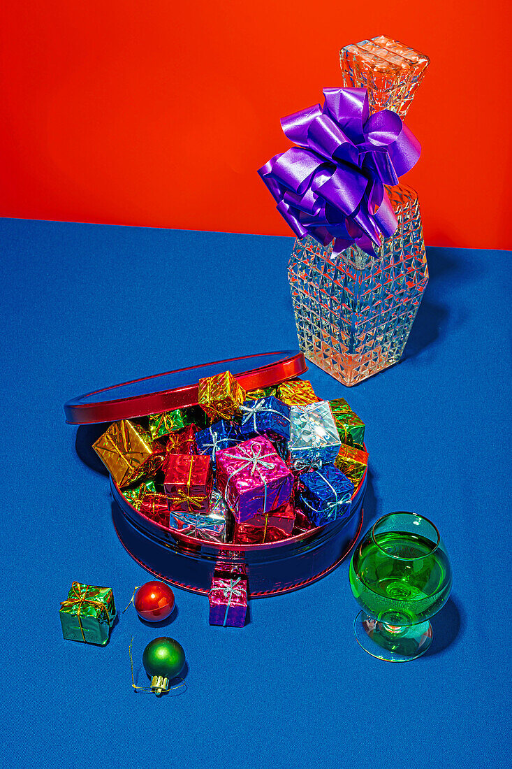 From above colorful Christmas gifts in a red tin and a sparkling vase with a purple bow on a vibrant blue table against a red background
