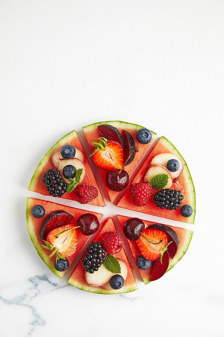 Top view of ingredients consisting of strawberry apple raspberry grapes blackberry plum blueberry peach placed on round cut slices on white surface while preparing watermelon pizza