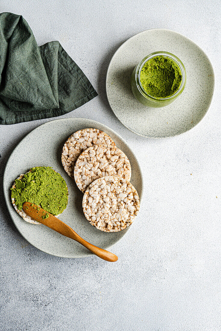 Top view of rice bread on a plate accompanied by a vibrant green spinach pesto pasta-sauce in a glass jar, set against gray backdrop near napkin