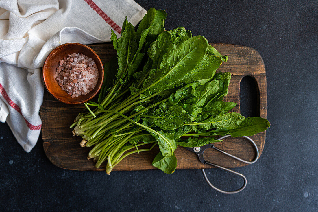 Top view of bunch of fresh arugula leaves alongside a bowl of pink salt and scissors on a rustic wooden cutting board, set against a dark tabletop with a striped towel