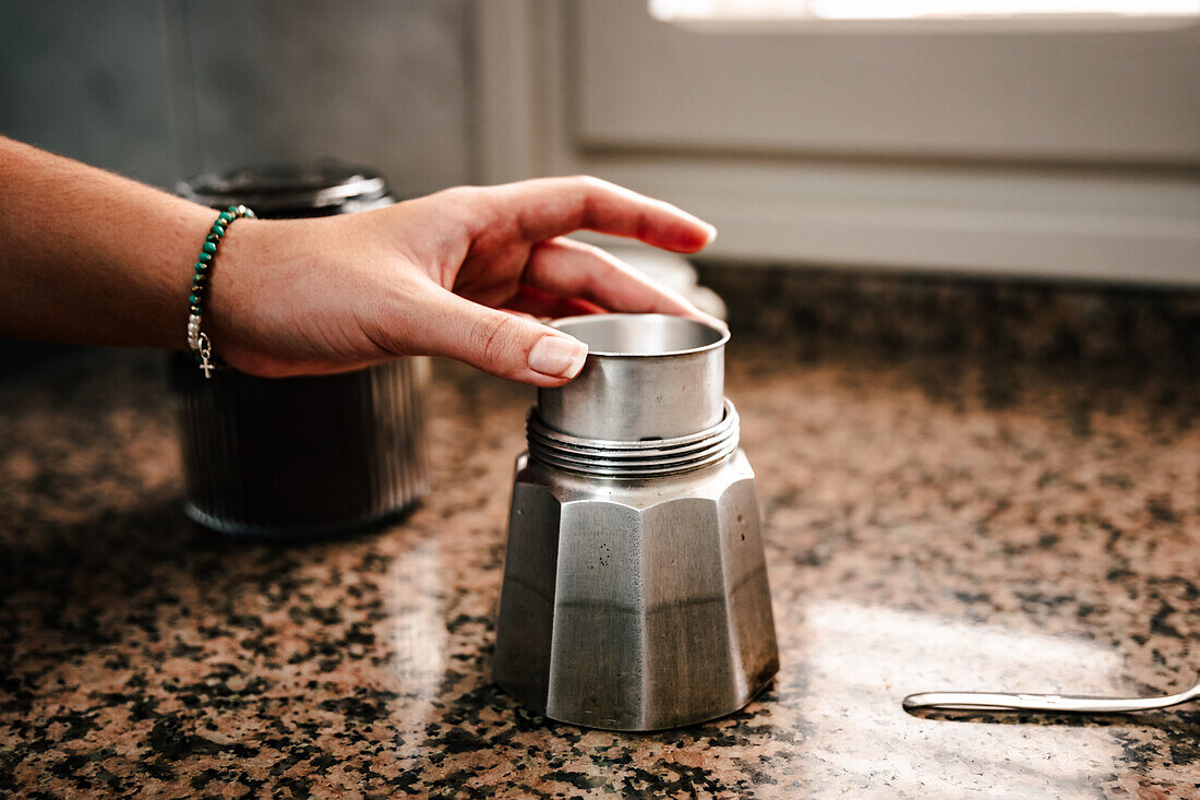 Close-up of anonymous hand assembling an Italian moka pot on a kitchen counter with blurred kitchenware in the background signifying a home coffee making process