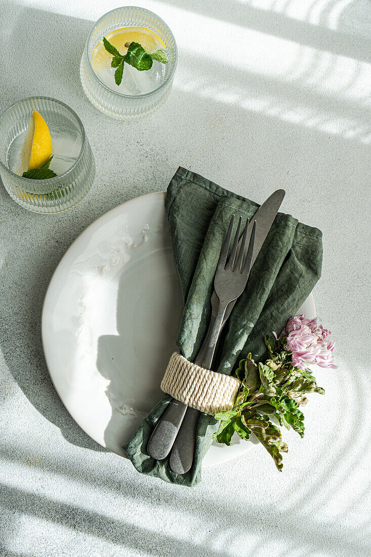 Ein elegant gedeckter Tisch mit weißen Tellern, in eine grüne Serviette eingewickeltem Besteck und natürlichen Dekorationen, wobei das Sonnenlicht zarte Schatten wirft