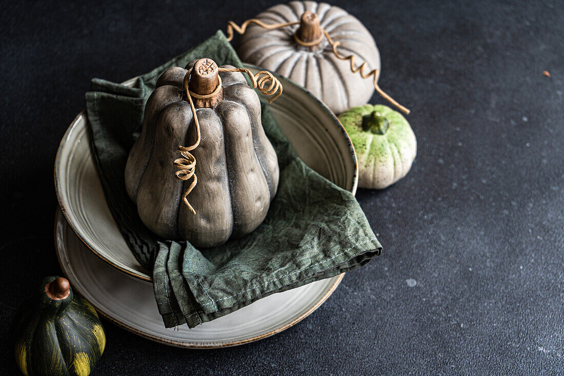 High angle of autumnal table setting with napkin and pale colored pumpkin placed on ceramic bowl near pale green pumpkins against dark surface