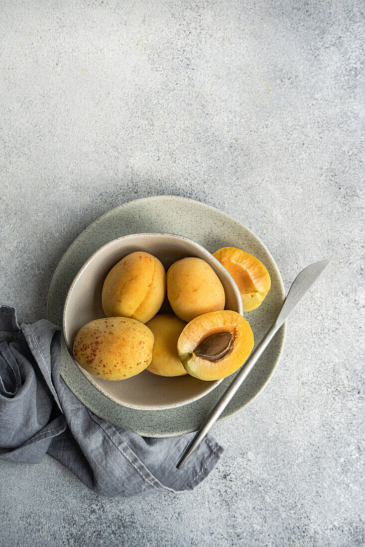 Draufsicht auf eine Keramikschale mit frischen reifen gelben Aprikosen auf blauem Tuch auf einem Betontisch mit halbierter Aprikose