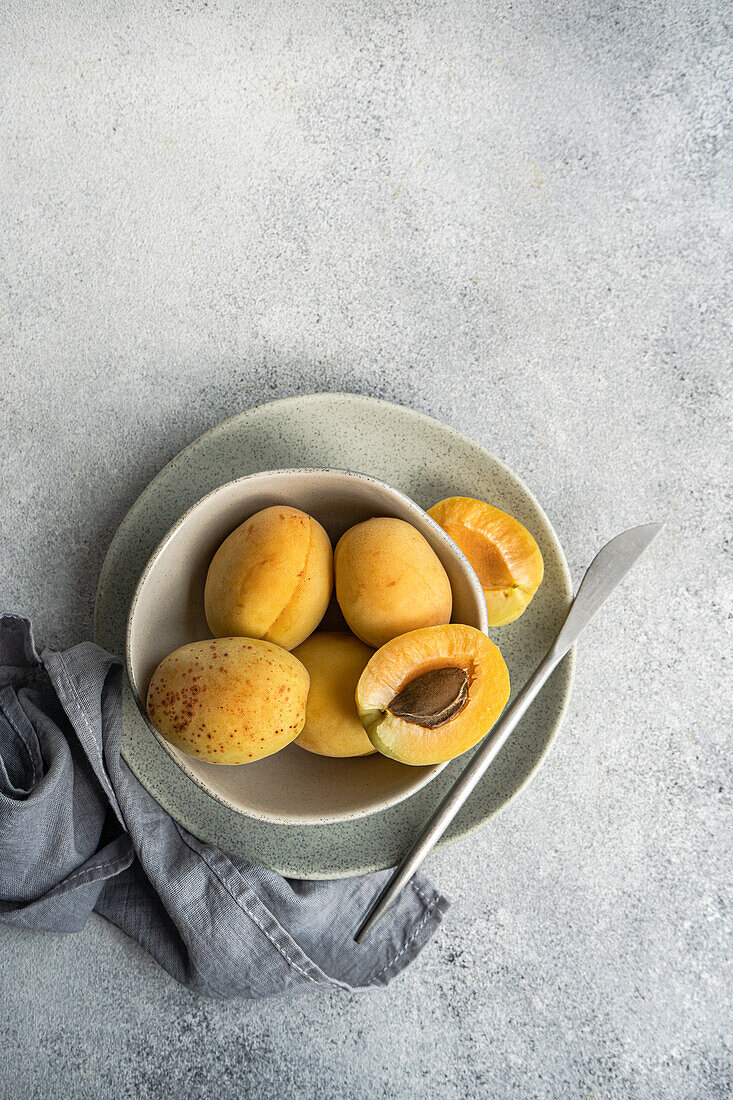 Top view of ceramic bowl with fresh ripe yellow apricots placed on blue cloth on concrete table with cut in half apricot