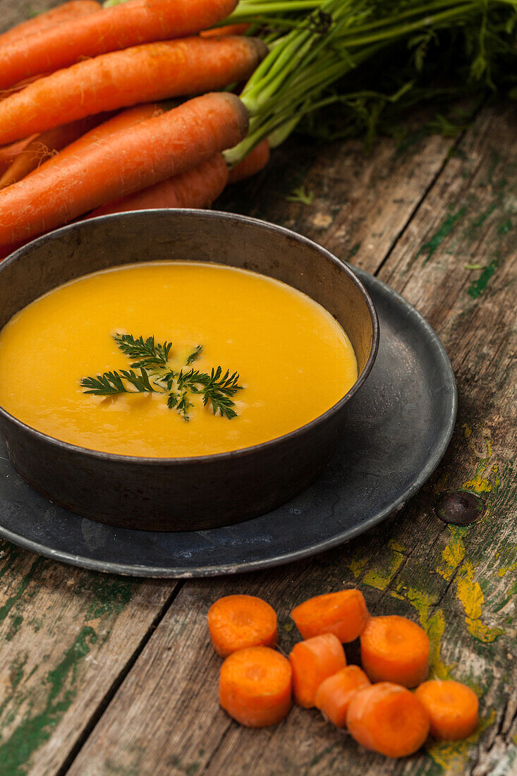 Rustic and homemade carrot soup served in a dark bowl, garnished with parsley, alongside fresh carrots with greens on a weathered wooden background.