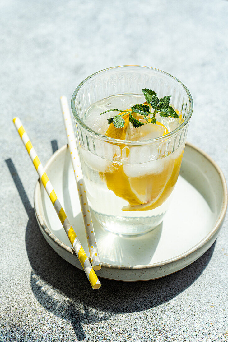 High angle of Summer cocktail with lemon vodka, slices of lemon and wild mint leaves with ice served on plate near straws on gray table