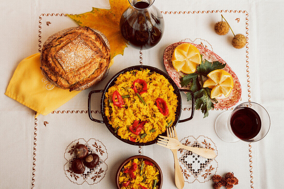 Overhead view of a traditional Spanish paella in a pan, served with bread, wine, and garnishes on a patterned tablecloth.