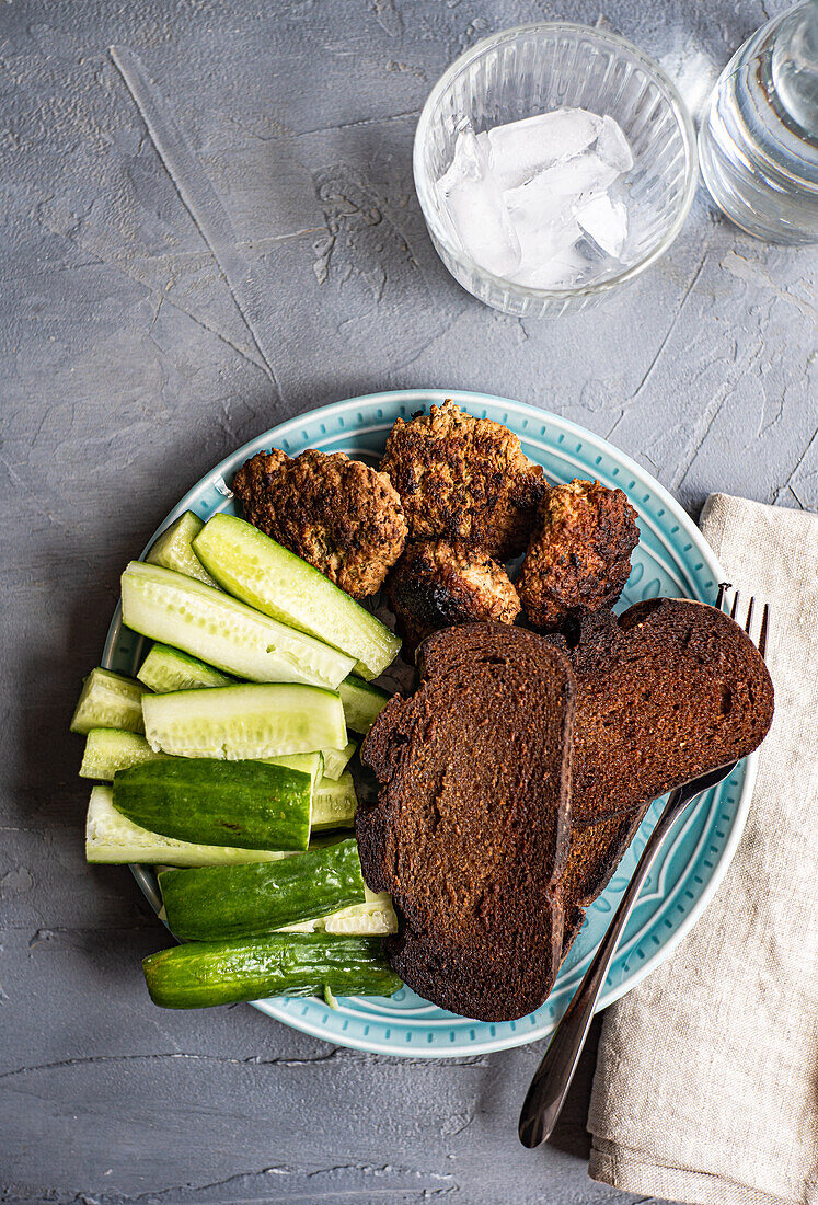Top view of fresh organic cucumber, slices of rye sour dough bread and meat cutlets served on plate near jar of water, glass, fork and napkin against gray background
