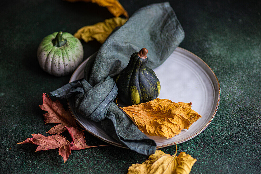 High angle of autumnal table setting with napkin, leaves and pumpkins placed on plate against dark surface