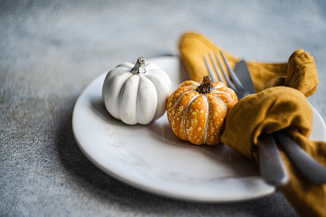 White ceramic plate with decorative pumpkins with silver fork and knife, wrapped in a mustard-colored napkin, placed on a textured gray concrete surface