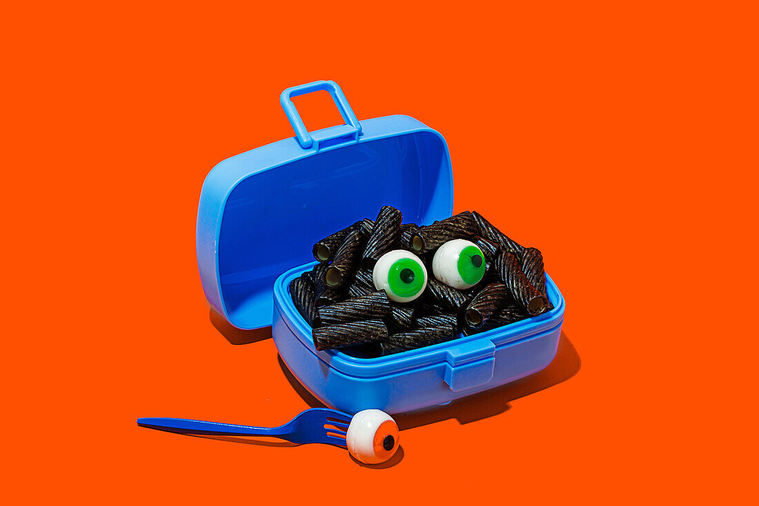 Horror lunch with black pasta and eyes in lunchbox placed on orange background near fork with eye