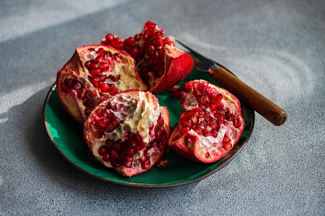 From above fresh pomegranate pieces with vibrant red seeds on a green ceramic plate set against a textured grey concrete background