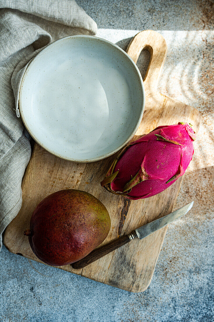 A vibrant pink dragonfruit alongside a ripe mango, a wooden cutting board, and a ceramic bowl on a textured surface.
