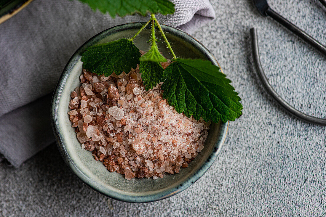 Nahaufnahme von oben eines veganen Kochgeschirrs mit einer Schale mit rosa Himalaya-Salz, garniert mit einem frischen Brennnesselblatt auf einem grauen Hintergrund mit einer Schere in der Nähe