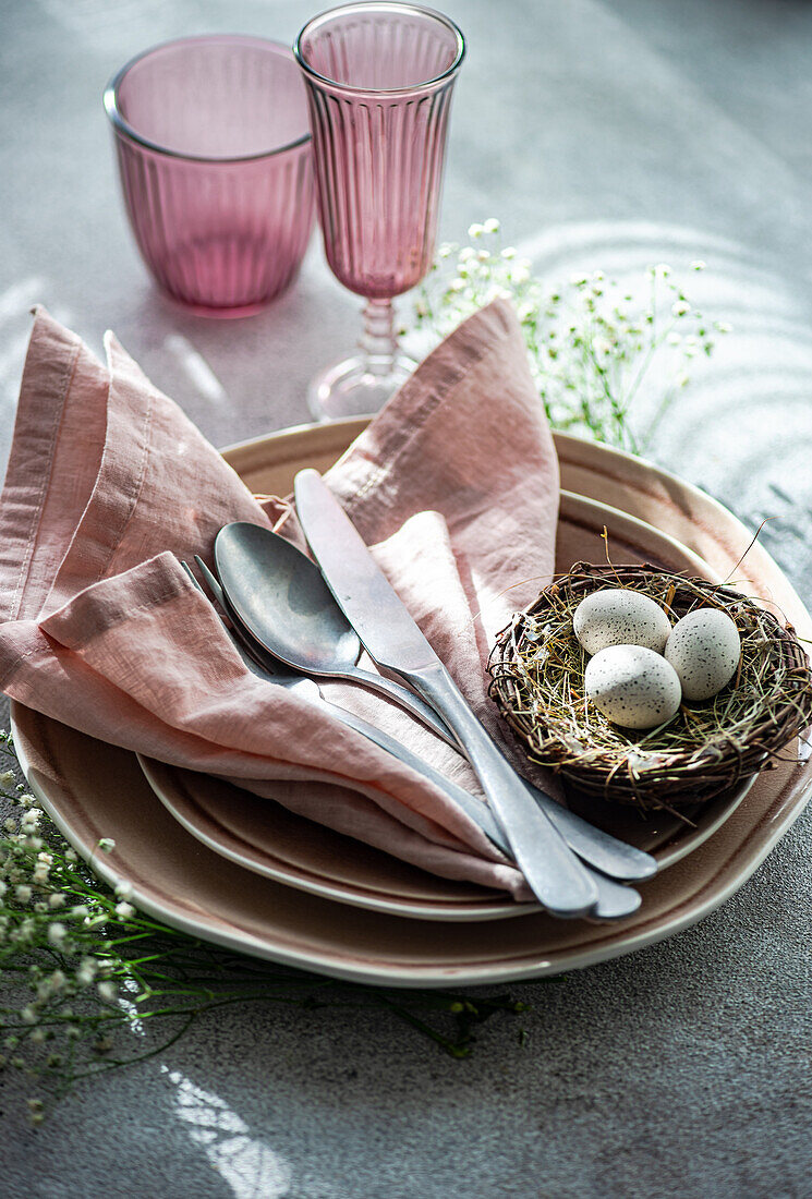 An elegant Easter dining setup featuring a nest with speckled eggs, pink napkins, and matching colored glassware on a textured surface.