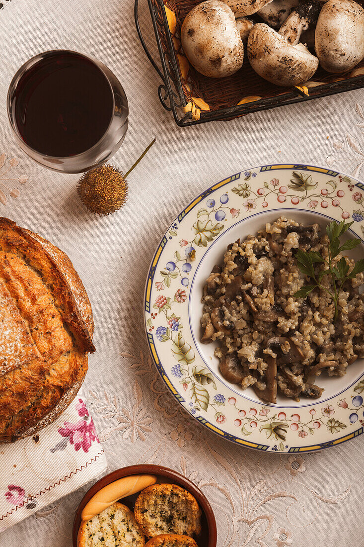 Blick von oben auf einen rustikal gedeckten Tisch mit Pilzrisotto, frisch gebackenem Brot und einem Glas Rotwein