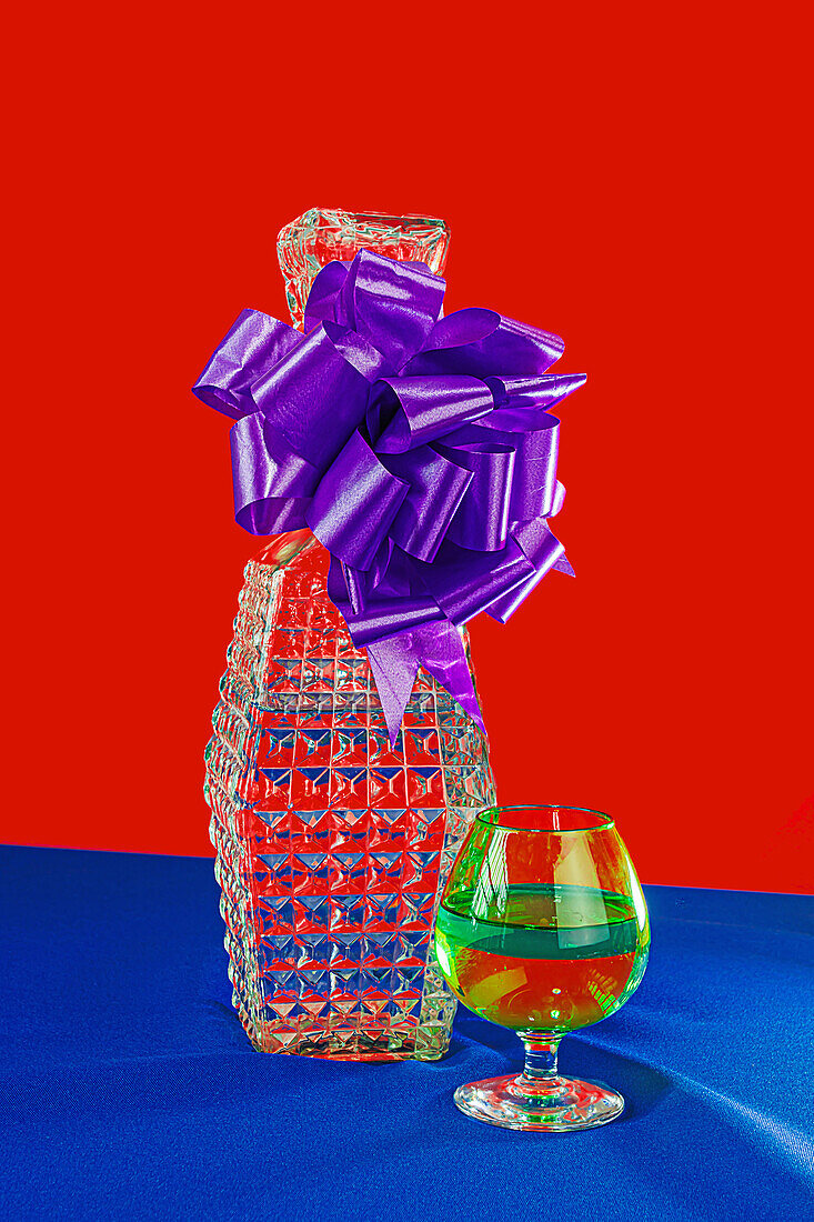 A festive crystal vase adorned with a large purple bow and colorful ribbons next to a multicolored glass on a blue cloth