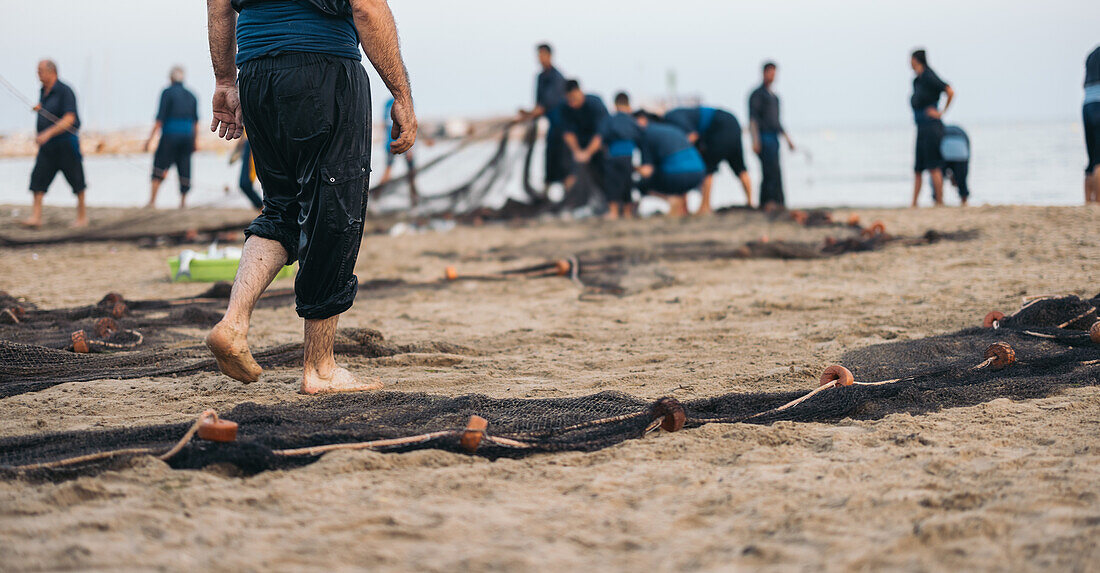 Rückenansicht eines anonymen Mannes in nasser Kleidung beim Trocknen eines Fischernetzes am Sandstrand vor einer unscharfen Gruppe von Fischern mit ihrem Fang