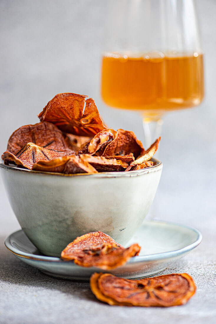Slices of dried apples served in ceramic bowl on plate near glass of juice against blurred gray background
