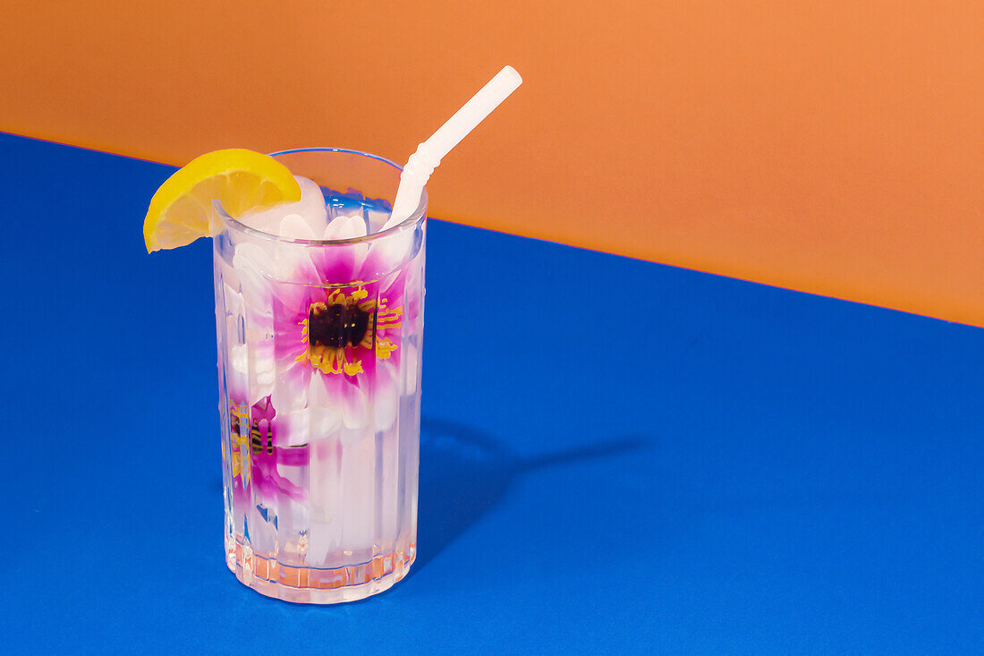 Transparent glass of refreshing cold drink decorated with pink flowers and straw placed on blue surface against bright orange wall