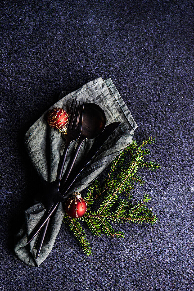 Von oben gedeckter Tisch für ein Weihnachtsessen mit dunklem, modernem Besteck, eingewickelt in eine grüne Serviette mit Tannenbaumblättern und Dekor auf einem Betontisch