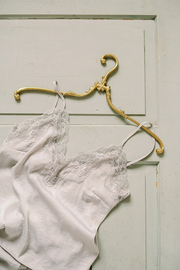 Weißes Seidentop mit Spitzenverzierungen neben goldenem eleganten Kleiderbügel auf Vintage-Oberfläche