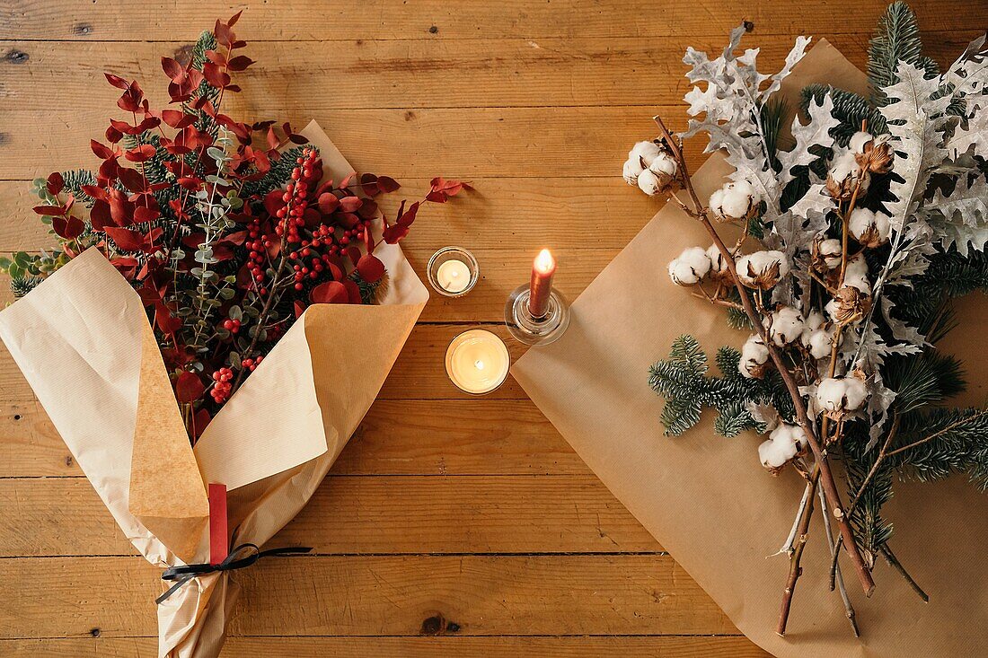 Festlicher Weihnachtsstrauß mit Baumwoll-, Tannen- und Eukalyptuszweigen und leuchtend roten Zweigen mit Beeren auf einem Holztisch mit Kerzen im Raum von oben