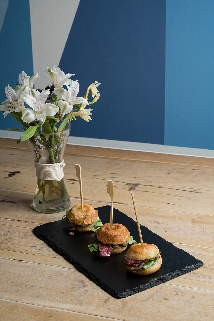 Frische, appetitliche Mini-Burger, serviert auf einem schwarzen Brett neben einer Vase mit blühenden Lilien an einer blauen Wand