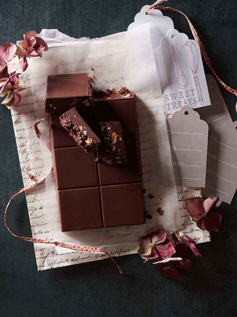 Draufsicht auf eine leckere süße Schokoladentafel auf Geschenkpapier mit getrockneten Blumen und Anhängern auf dunklem Hintergrund im Zimmer