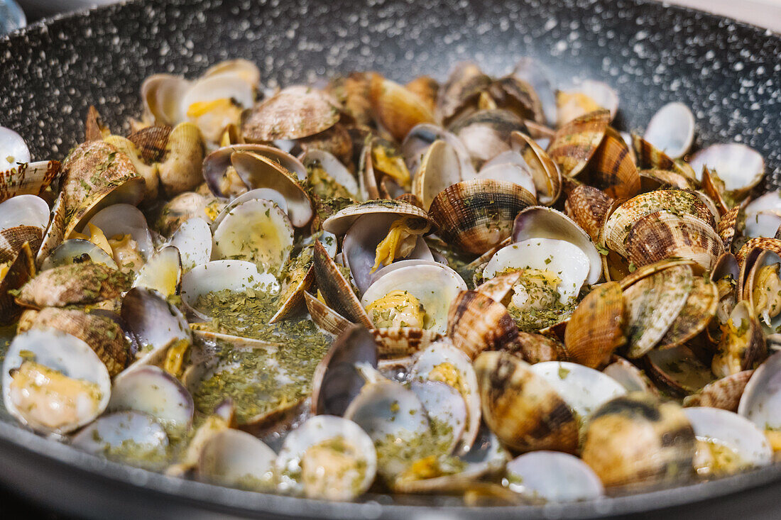 Haufen schmackhafter Mollusken mit Schalen, die in einer heißen schwarzen Bratpfanne auf einem Elektroherd in einer hellen Küche zu Hause gebraten werden