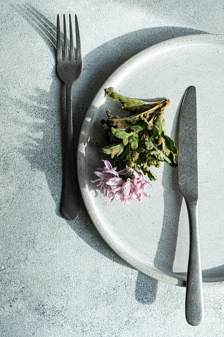 A top-down view of a white plate with pink flower alongside a fork and knife on a textured gray surface.