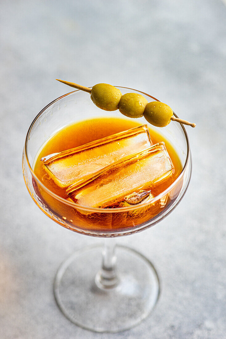 Hoher Winkel eines Glases mit einem alkoholischen Cocktail mit Whiskey und Sticks mit öligen Olivenscheiben auf grauem Hintergrund im Studio