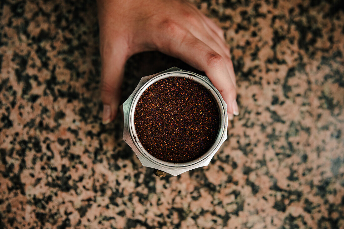 Top view of freshly ground coffee filled to the brim of stainless steel stovetop espresso maker with anonymous young person hand holding the base and a sealed coffee jar nearby on textured countertop